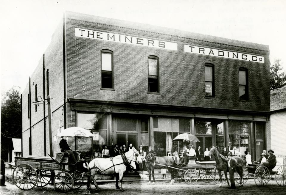 black and white photo of a large brick building with glass front and the words "The Miners Trading, Co" Many people and horse drawn carts line the street in front of it. 