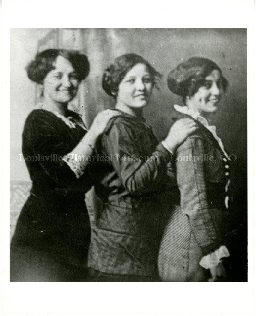 Three women standing sideways looking at camera with hands on each others shoulders.
