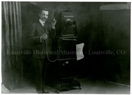 Man posing with large camera on stand with sign reading "smile please".