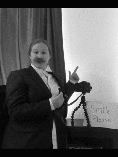 Woman dressed as man posing next to small camera on a tripod with a sign that reads, "smile please"