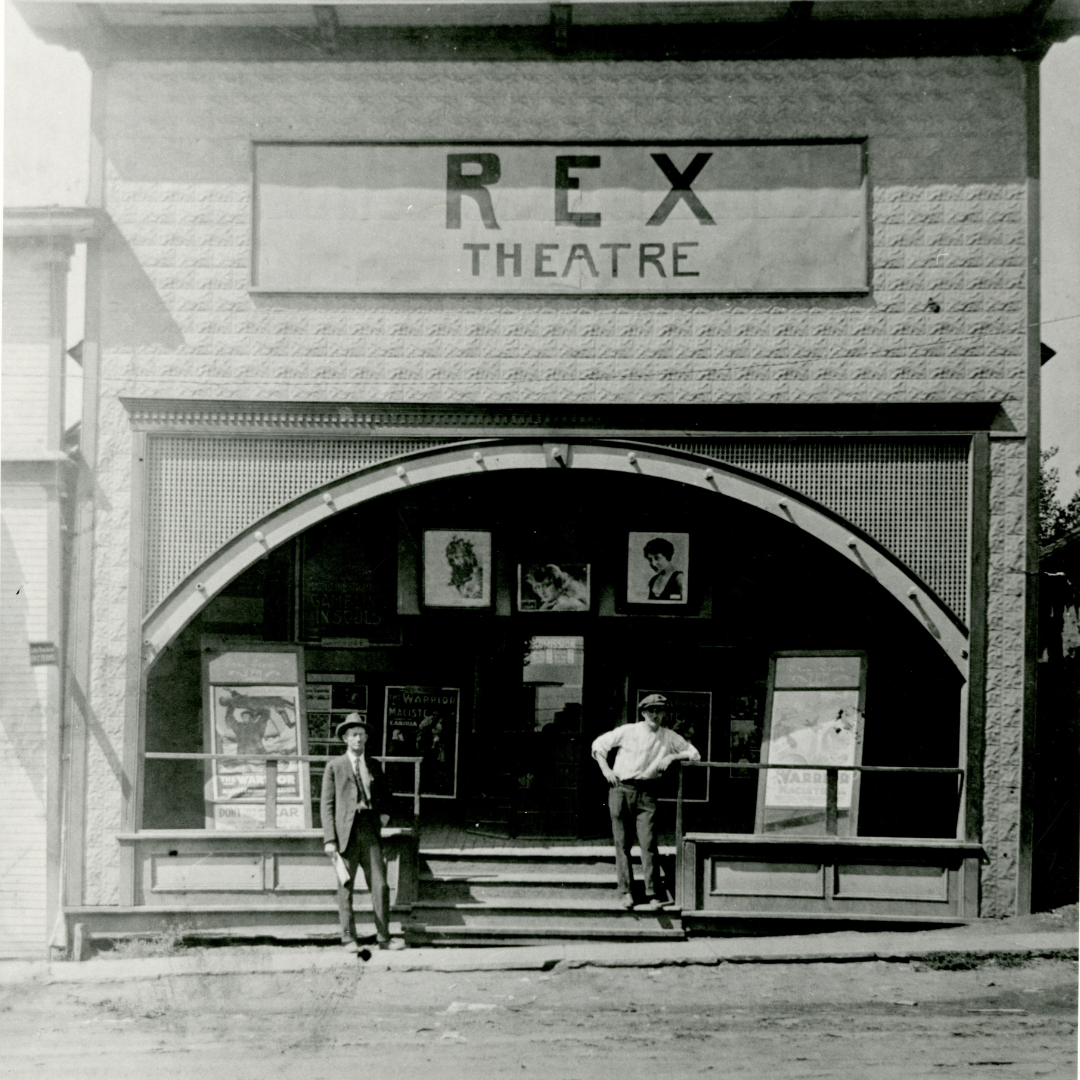Building that says Rex Theatre with movie posters and two men standing in front of it