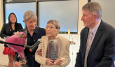Memory Delforge holds a plaque and speaks at a microphone, with her two adult children