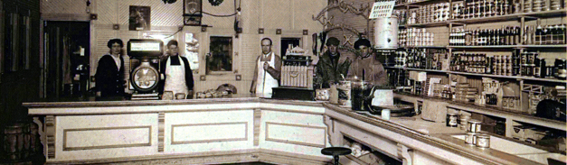 black and white photo of men behind a counter of a grocery store. Cans and bottles line the wall.