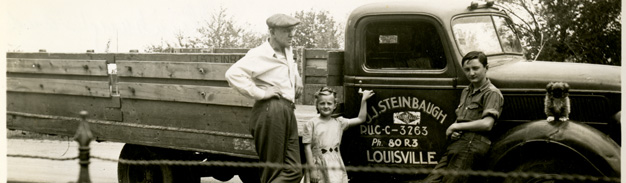 black and white photo of man, girl, boy, and a small dog standing next to an old pick up truck that reads, "J. J. Steinbaugh, Louisville"